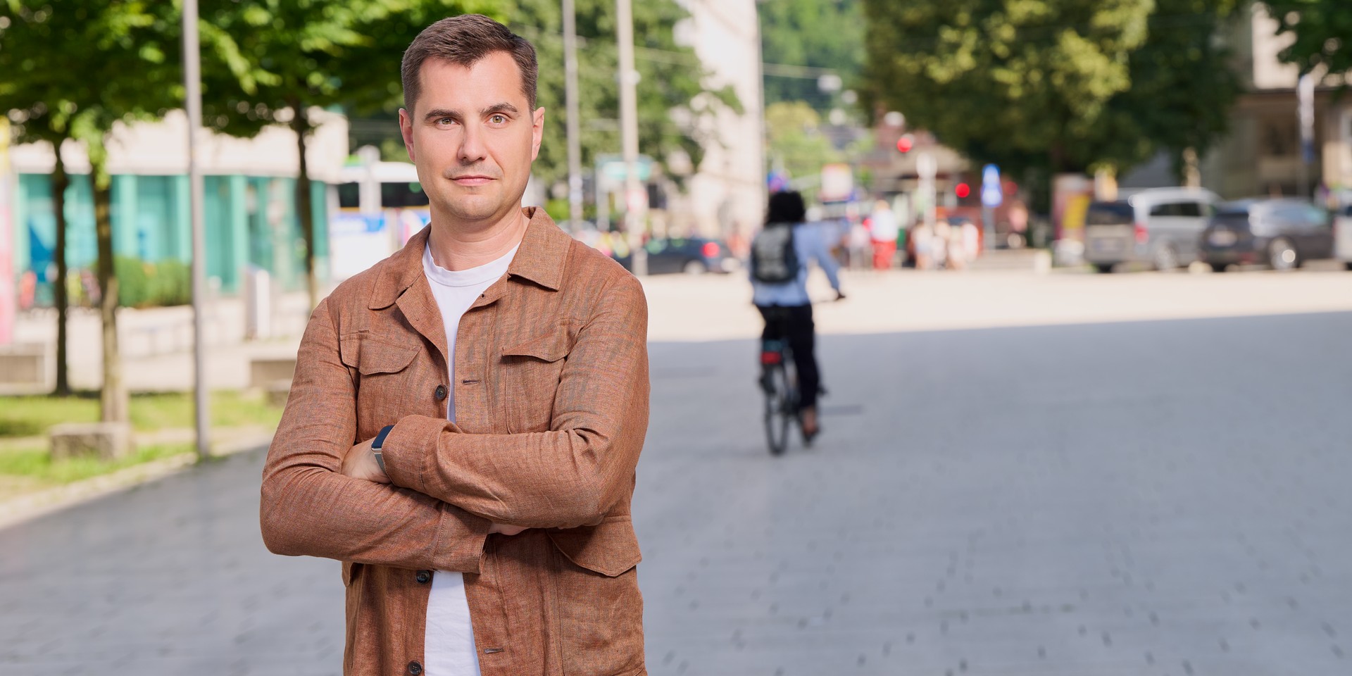 Mann mit verschränkten Armen steht in einer Stadt, Radfahrer im Hintergrund
