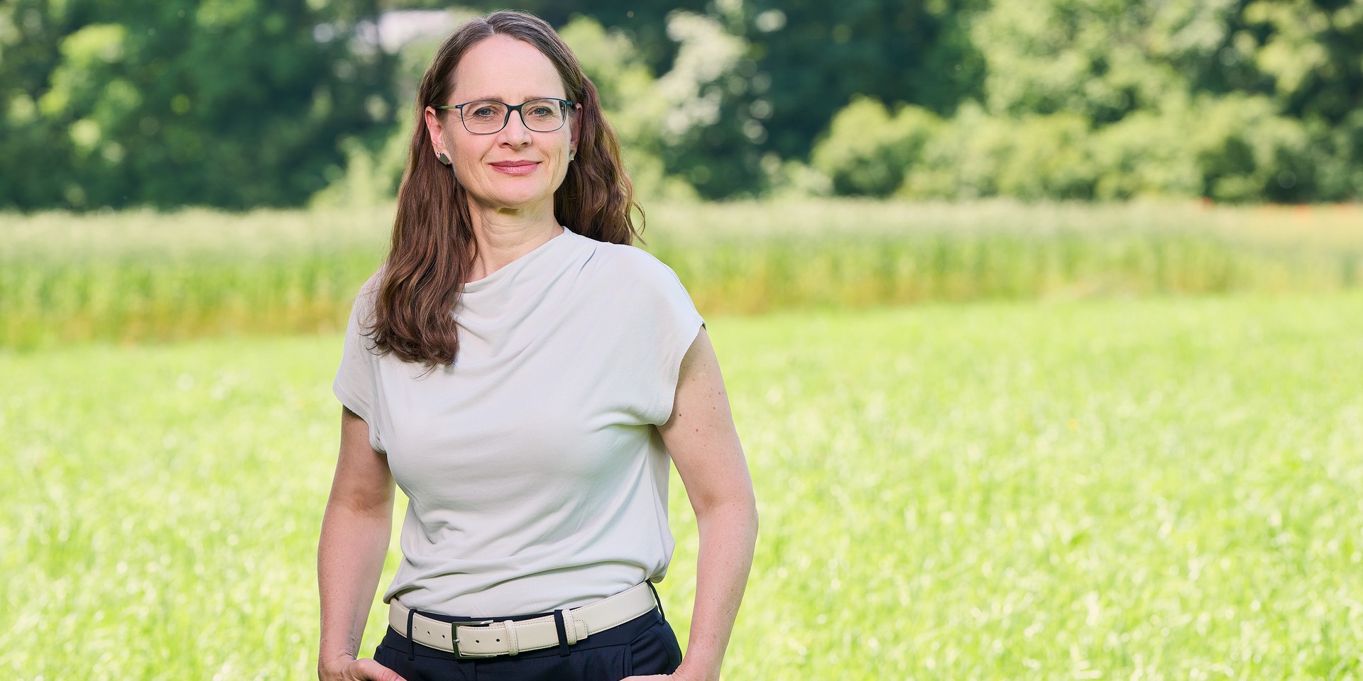 Frau mit Brille und braunen Haaren steht auf einer grünen Wiese vor Bäumen.